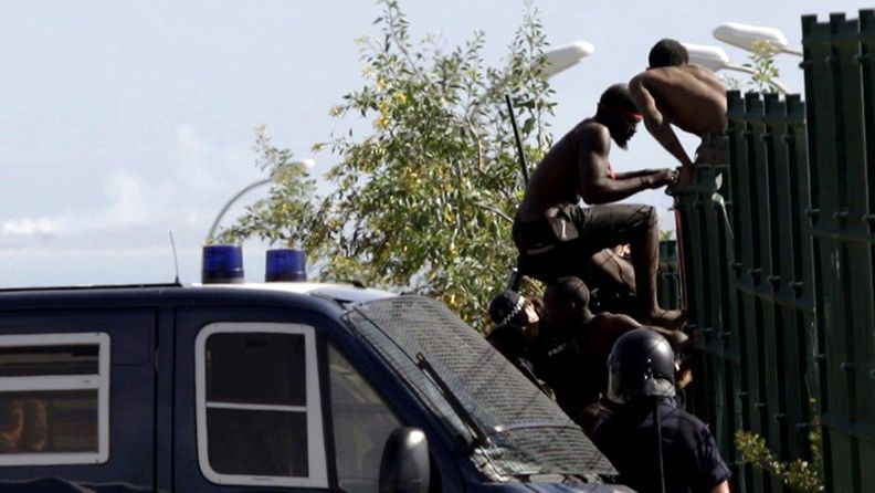 Siirtolaisia kiipeämässä Melillan kaupunkiin poliisiauton odottaessa.