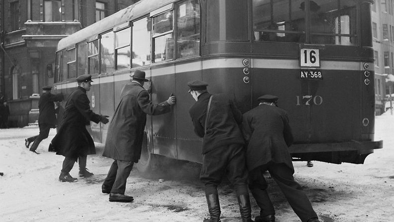 Helsingin Liikennelaitoksen sininen bussi poikittain märän lumen liukastuttamalla Erottajan mäellä. Kuljettajat ja matkustajat yrittävät kääntää bussia oikeaan suuntaan.