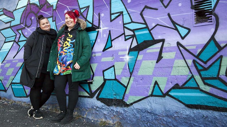 Sateenkaarinuorisotyöntekijät Mari-Katri Lempinen ja Emmi Erkkilä nojaavat graffitiseinään.