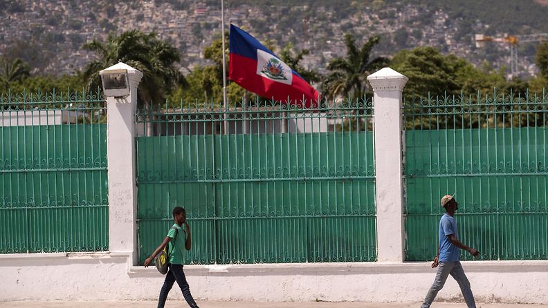 Haitilaiset kävelevät korkean aidan edustalla, jonka takana liehuu valtion lippu puolisalossa.