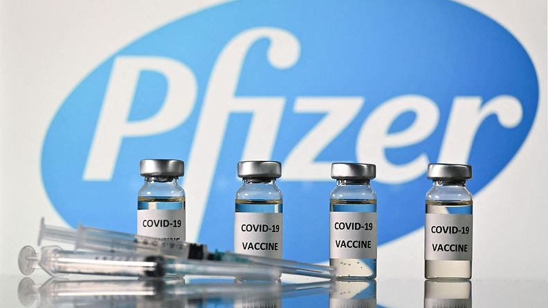Covid-19 rokoteputeleita rivissä ja ruiskut lepäävät pöydällä edessä. Kimmeltävien purkkien taustalla paistaa Pfizerin logo.