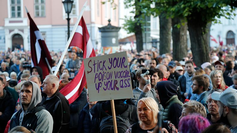Joukko mielenosoittajia. Latvian liput liehutvat ja nainen pitää kylttiä, jossa lukee "Stick your poison vaccine up your ass".