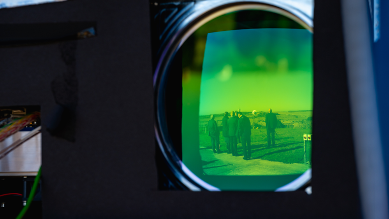 Lähikuvaa lasertykin laitteistosta. Vihreän linssin heijastuksessa näkyy ulkona seisovia ihmisiä.