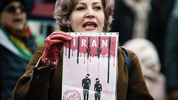 Mieltään osoittava nainen pitelee kylttiä, jossa on piirroskuva hirtetyistä ihmisistä ja tekstit "IRAN" ja "STOP EXECUTIONS"