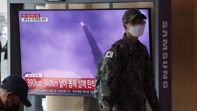 Harmaaseen maastopukuun pukeutunut sotilas kävelee maski kasvoillaan tv:n ohi, jonka ruudulla näkyy uutinen laukaistusta ohjuksesta.