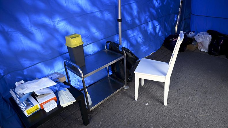 Sinisen teltan sisällä on valkoinen tuoli ja pöytiä, joiden päältä löytyy huumeiden käyttöön tarvittavat välineet.