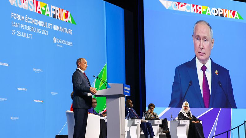Putin puhuu puhujanpöntöllä huippukokouksessa, suuret näytöt taustalla heijastavat kokouksen logoa ja Putinin kuvaa.