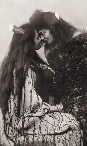 AOP Maorien tapa tervehtiä painamalla nenää. Toisella tytöllä on pellavaviitta ja hiuksissaan huia-sulka, arvomerkki, ja toisella pellavapohjan höyhenistä tehty viitta. 1800-luvun valokuva.